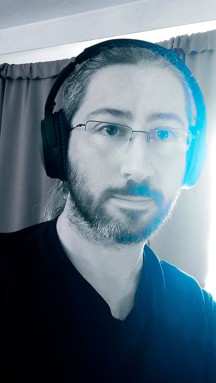 autoportrait de Smalt en noir et blanc. Il porte un t-shirt col en V, une barbe taillée court, des lunettes et un casque sur les oreilles.
